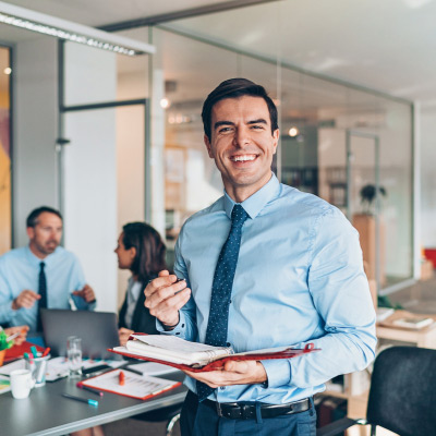 Manager souriant tenant un carnet tout en discutant avec son équipe en arrière-plan dans un bureau moderne